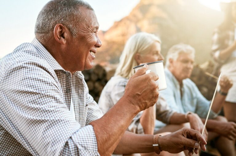 Entspannter Senior beim Camping mit Freunden, der eine Pause einlegt und eine Tasse Kaffee trinkt, während er den Ruhestand genießt und sich draußen in der Natur gesund hält. Lächelnder Mann im Ruhestand auf einem Wellness-Rückzugsort.